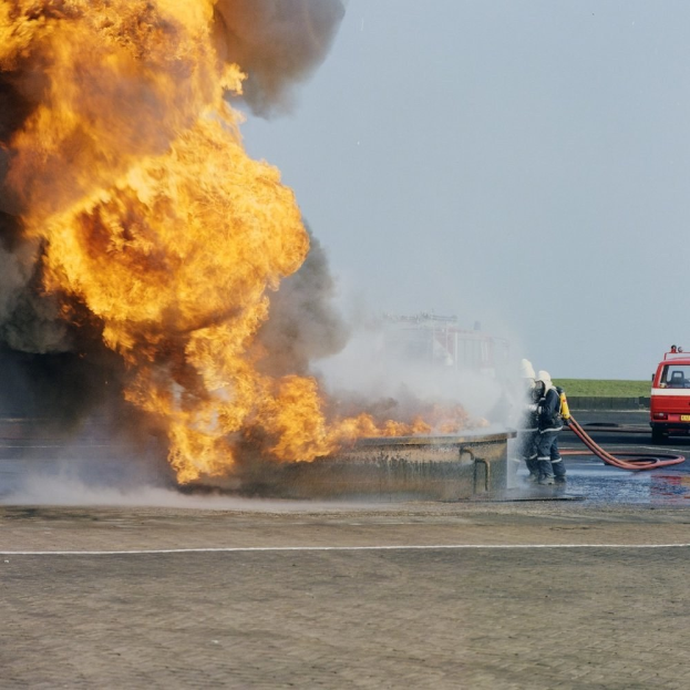 Ein Feuerwehrauto brennt am Straßenrand mit zwei Personen in Helmen und Schläuchen, ein Fahrzeug im Hintergrund und der Himmel sichtbar.