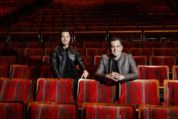 Zwei Männer in schwarzen Jacken lächeln, während sie auf roten Auditoriumsstühlen während einer Vorstellung von *Dancing with the Stars* sitzen.