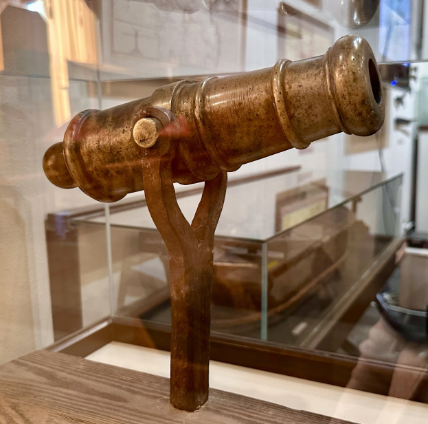 Alte Messingkanone in einer Glasvitrine mit einem Holzuntergrund, vor gerahmten Wandausstellungen in einem Museums Setting.