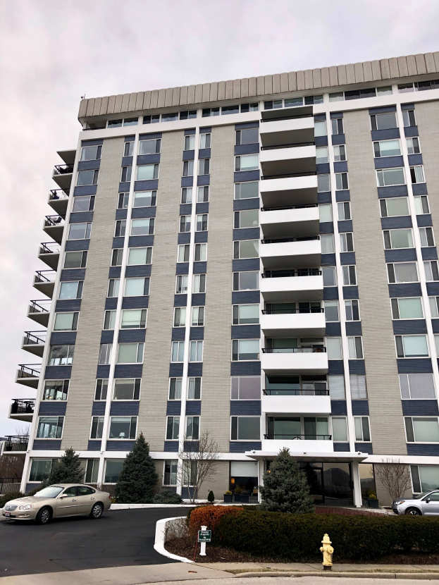 Hohes Wohngebäude mit zahlreichen Fenstern, einem roten Feuerhydranten im Vordergrund, Fahrzeugen auf der Straße, Grünflächen einschließlich Pflanzen und Bäumen sowie einem bewölkten Himmel im Hintergrund.