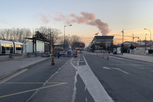 Eine Stadtstraße mit Verkehrskegeln, einem Bus, Laternenmasten, Bäumen, Gebäuden, Kränen und Rauch, der aus einem fernen Gebäude aufsteigt, vor einem Himmel.