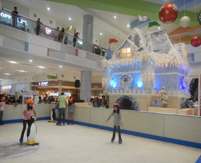 Eine Gruppe von Menschen, die auf einer Eisbahn in einem Einkaufszentrum Schlittschuh laufen, umgeben von einem hausähnlichen Aufbau, der mit Lichtern und festlichen Dekorationen geschmückt ist, mit Menschen, die auf dem Boden, Geländern, Säulen und Tafeln mit Text stehen, unter einer Decke mit Lichtern.