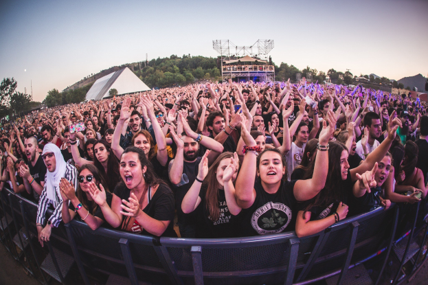Eine große Menschenmenge auf einem Musikfestival mit erhobenen Händen, die vor einem Geländer stehen, mit Bäumen, Gebäuden und einem klaren blauen Himmel im Hintergrund.