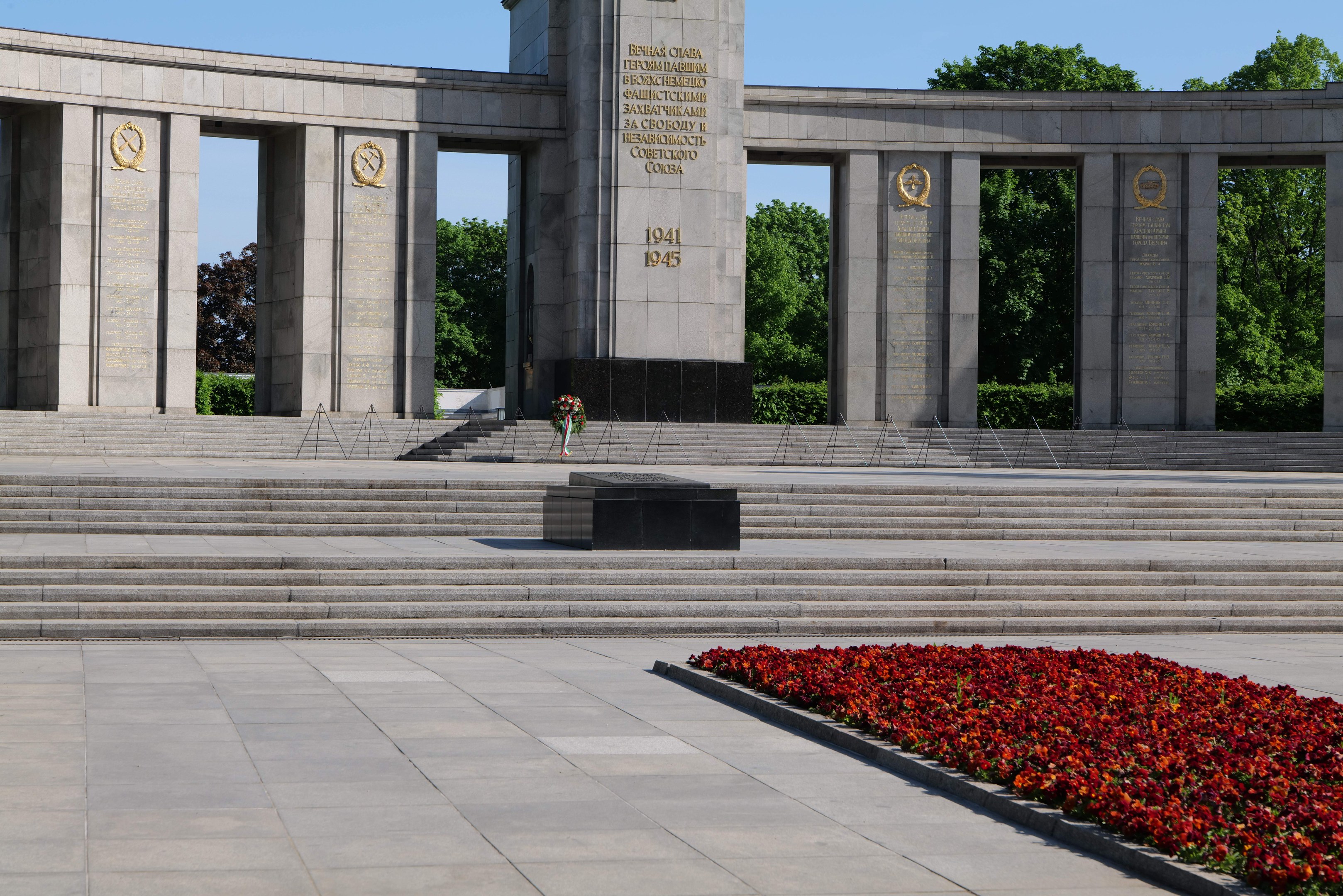 Ein Gedenkmonument in einem Park mit roten Blumen davor, Inschrift auf seinen Wänden und Stufen, umgeben von Bäumen unter einem klaren blauen Himmel.