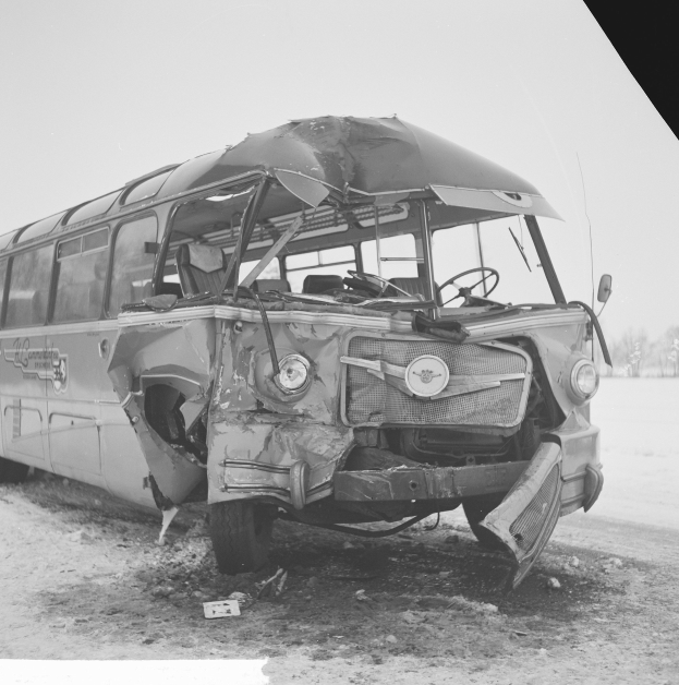 Schwarz-weiß-Foto eines schwerbeschädigten Busses mit eingedellter Front und zersplitterten Fenstern an der Seite einer Straße, umgeben von Bäumen unter einem bewölkten Himmel.