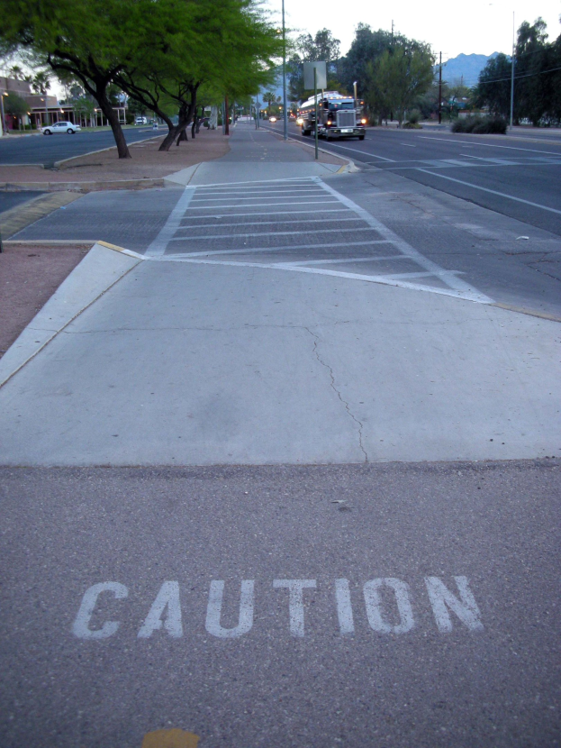 Eine Stadtstraße mit fahrenden Fahrzeugen, ein Gehweg mit einer Warnschildern in der Nähe eines Baumes, Strommasten, Straßenlaternen, Bauzäune, Grünflächen, Gebäude, ferne Berge und einen klaren blauen Himmel.