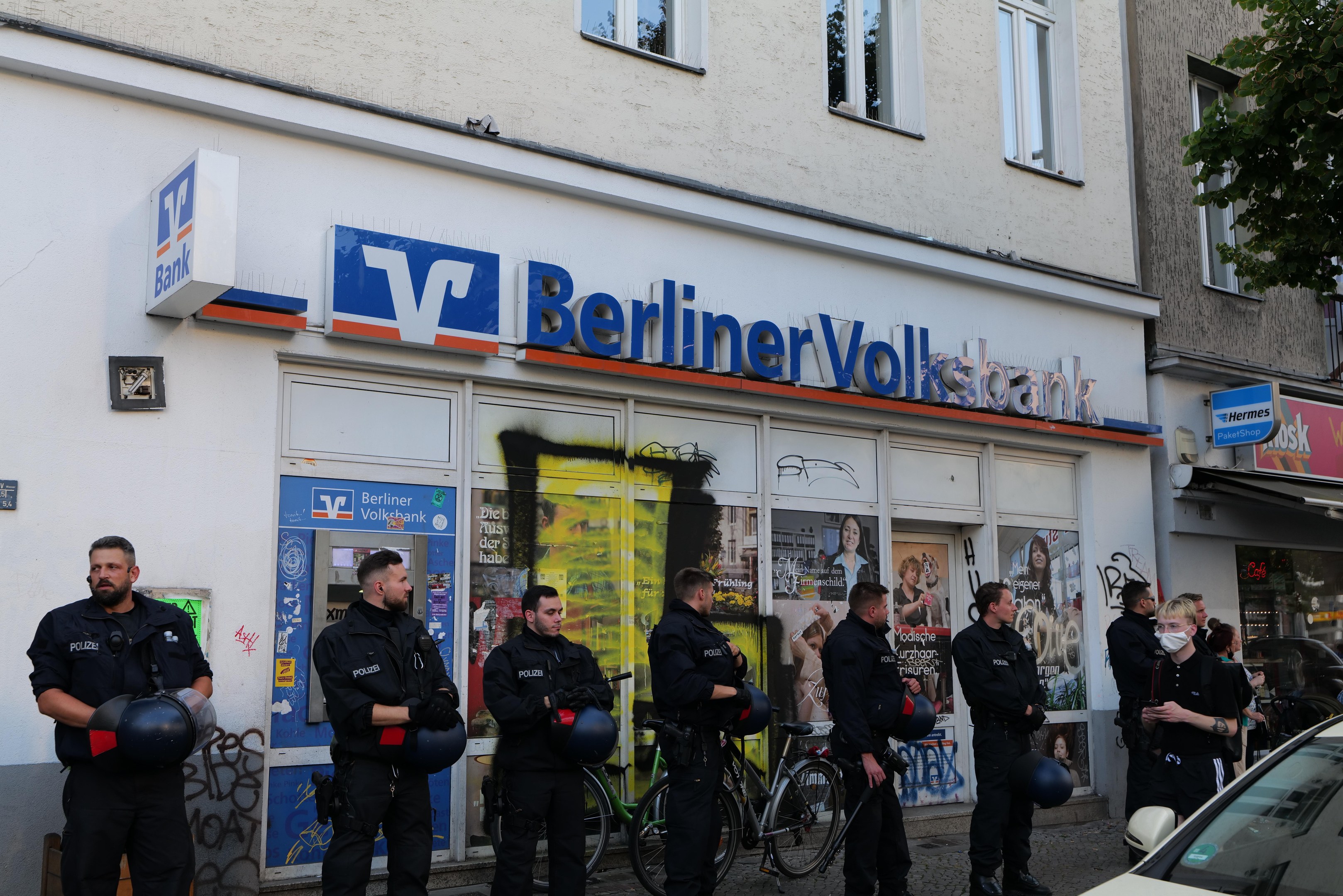 Gruppe von Polizisten vor einer Berliner Volksbank, einige mit Helmen, daneben ein Auto und ein Baum, Gebäude und Schilder im Hintergrund.