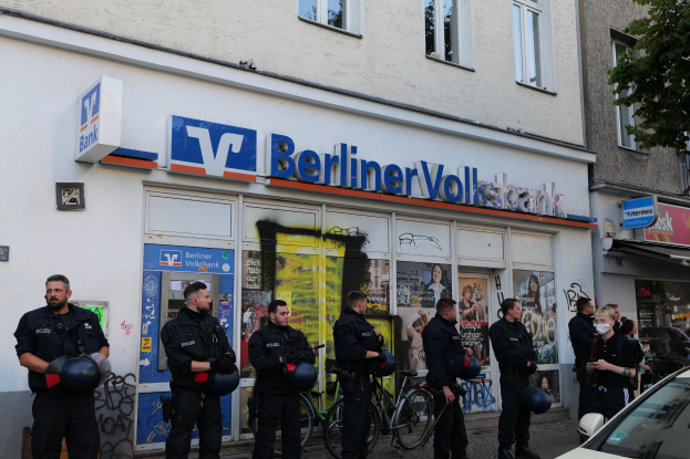 Gruppe von Polizisten vor einer Berliner Volksbank, einige mit Helmen, daneben ein Auto und ein Baum, Gebäude und Schilder im Hintergrund.