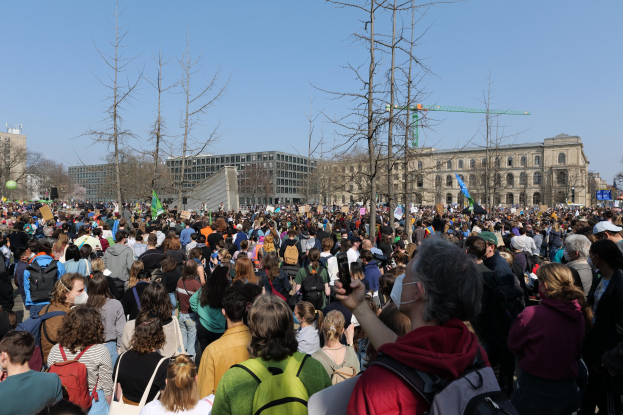 Eine große Menge Menschen mit Schildern und Taschen steht vor einem Gebäude mit Bäumen und einem klaren Himmel, was auf eine Klimawandel-Demonstration hinweist.