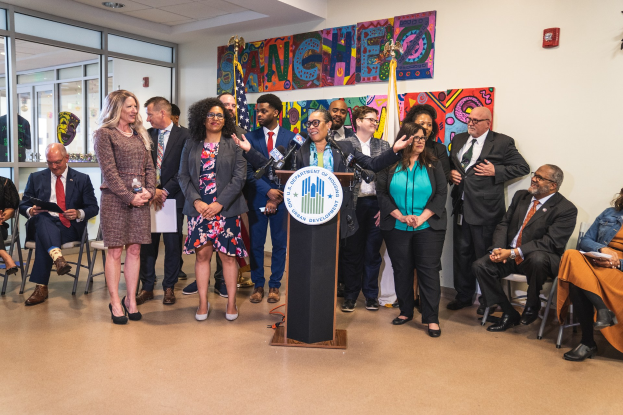 Gruppe von Menschen in einem Raum mit einem Podium, Mikrofon und einer Tafel mit der Aufschrift "Sancho City Councilman Joins City Council", umgeben von Stühlen, Flaggen, einem Feuerlöscher und Deckenleuchten.