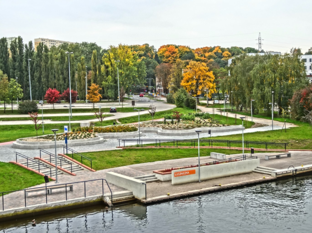Ein Fluss fließt durch einen grünen Park neben hohen Gebäuden, mit Bäumen, Straßeninfrastruktur, Fahrzeugen und einem bewölkten Himmel.