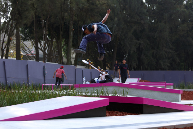 Ein Skateboarder, der einen Trick in der Luft auf einer Rampe ausführt, umgeben von Zuschauern mit Skateboards und verstreuten persönlichen Gegenständen vor einem Hintergrund aus Bäumen, Gebäuden und einem klaren Himmel.