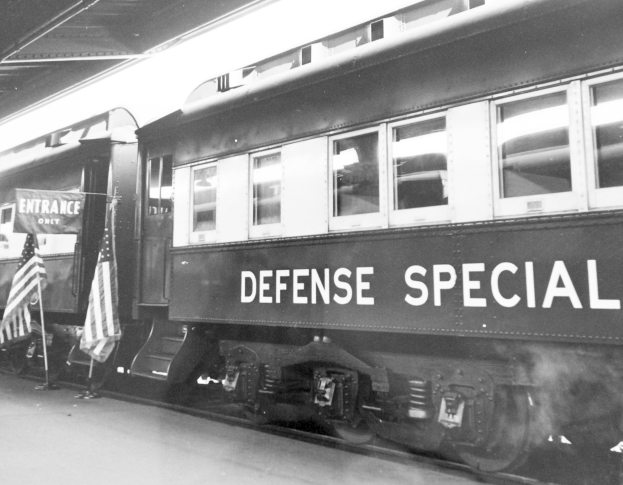 Schwarzes und weißes Foto eines Zuges mit der Aufschrift "Defense Special" auf Gleisen mit zwei Fahnen links und einer Decke oben.