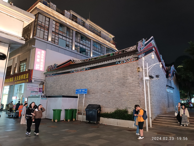 Menschen, die nachts eine Straße mit Gebäuden, Straßenlaternen, Schildern, Bäumen und Mülltonnen entlanggehen, tragen einige Taschen.