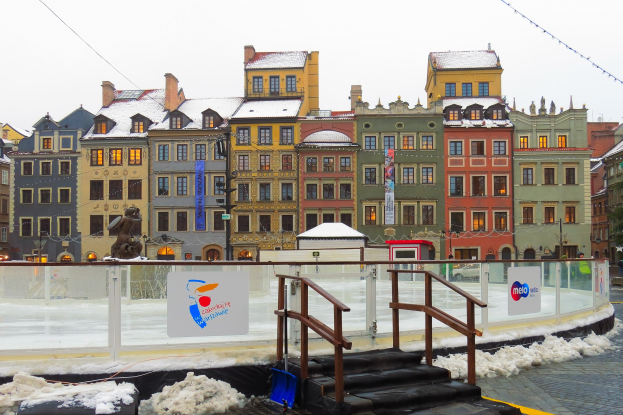 Eislaufbahn in einem Stadtplatz umgeben von Gebäuden, Treppen, Geländern, Informationsschildern, schneebedecktem Boden, Laternen, Bäumen und einem Himmel als Hintergrund.