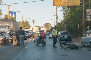 Eine Gruppe von Menschen umringt ein verunglücktes Motorrad am Straßenrand mit mehreren Fahrzeugen, darunter ein Lastwagen, und einem Hintergrund aus Bäumen, Pfählen, Lampen und Schildern unter dem Himmel.