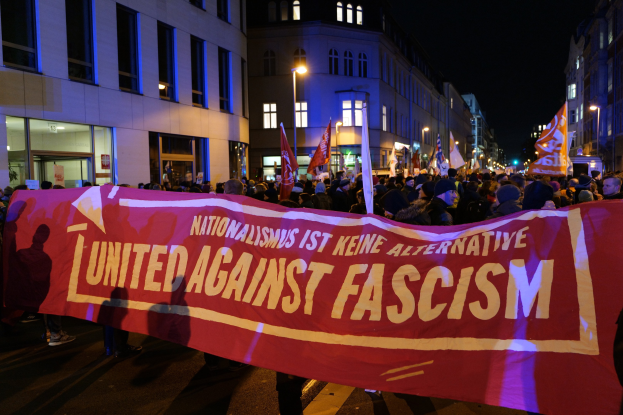 Eine Gruppe von Menschen marschiert auf einer Stadtstraße, die ein Banner mit der Aufschrift 'Einheit gegen Faschismus' trägt, mit Gebäuden, Straßenlaternen und Schildern im Hintergrund.