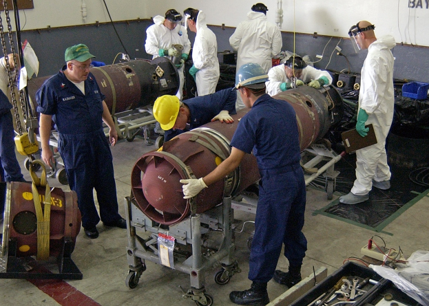 Eine Gruppe von Männern in Helmen und Handschuhen entschärft eine große Bombe in einer Fabrik mit Maschinen und einer textbedeckten Wand im Hintergrund.