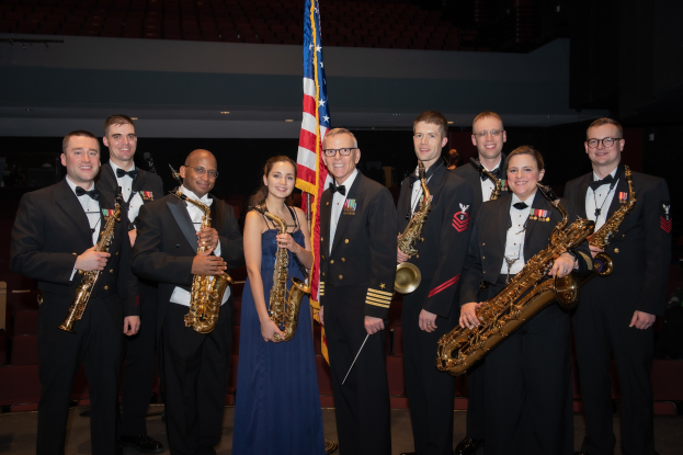 Gruppe von Menschen in Militäruniformen, die lächeln und Saxophone halten, mit einer Fahne und Stühlen im Hintergrund.