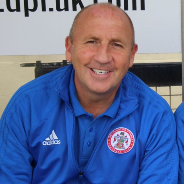 Zwei lachende Männer in einem Raum, einer in einer blauen Jacke, wahrscheinlich der Trainer von Accrington Stanley Football Club, mit einer Tafel an der Wand dahinter.