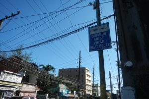 Stadtstraße mit fahrenden Autos, Strommäste mit Drähten, Gebäude, Bäume und Namenschilder, mit einem "Fahrer müssen Sitzgürtel tragen"-Schild an einem Strommast und einem sichtbaren Himmel im Hintergrund.