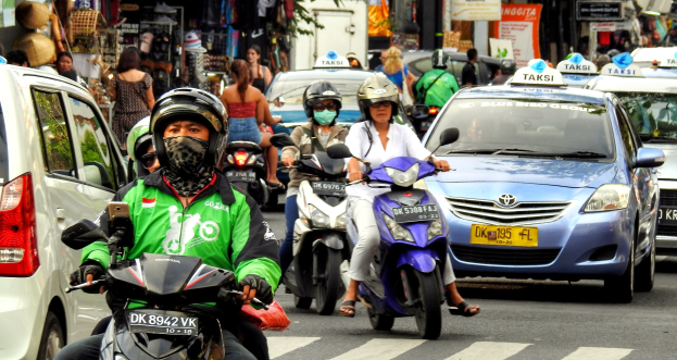 Eine Gruppe von Menschen auf Motorrädern fährt eine belebte Straße entlang mit Autos und anderen Fahrzeugen, einige Fahrer tragen Helme, mit Gebäuden, Bäumen, Schildern und Geschäften im Hintergrund.