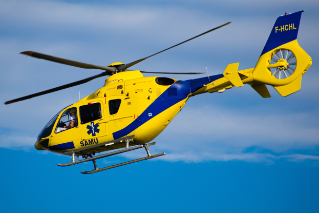 Gelb-blauer Hubschrauber mit "Samu Air Ambulance Service"-Text, der durch einen blauen Himmel mit Wolken fliegt.