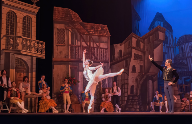 Zwei Tänzer performen auf einer Bühne im Royal Opera House in London, mit einem Publikum im Vordergrund und einem Bildschirm mit Gebäudeabbildungen im Hintergrund.