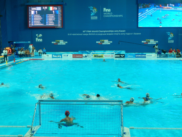 Eine Gruppe von Menschen spielt Wasserball in einem große Pool mit einem Netz in der Mitte, Zuschauern auf der linken Seite und einer Anzeigetafel mit Text und Logos im Hintergrund bei den FINA-Weltmeisterschaften 2016 in Kazan.