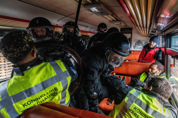 Polizeibeamte in Einsatzausrüstung auf einem Bus sitzend, mit einer Person in der Mitte, sichtbaren Fenstern auf der rechten Seite und einem Plakat an der Wand im Hintergrund.