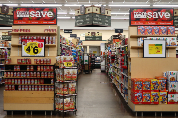 Ein Lebensmittelgeschäft mit Regalen und Boden voller verschiedener Lebensmittel, beleuchtet von Deckenlampen und mit Informationsschildern; eine Person steht im Hintergrund.