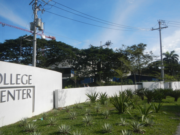 Eintritt in ein College-Center mit einem "College Center"-Schild an der Wand, umgeben von Pflanzen, einem Weg, Strommasten, Bäumen, Gebäuden, einem Kran und einem bewölkten Himmel.