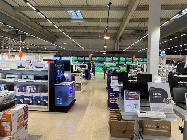 Ein großer Raum in einem Walmart-Geschäft voller Elektronik, die in Regalen angeordnet ist, einschließlich Computer und Drucker, mit Menschen auf dem Boden, einer Säule, Schildern, Deckenleuchten und Deckenventilatoren.