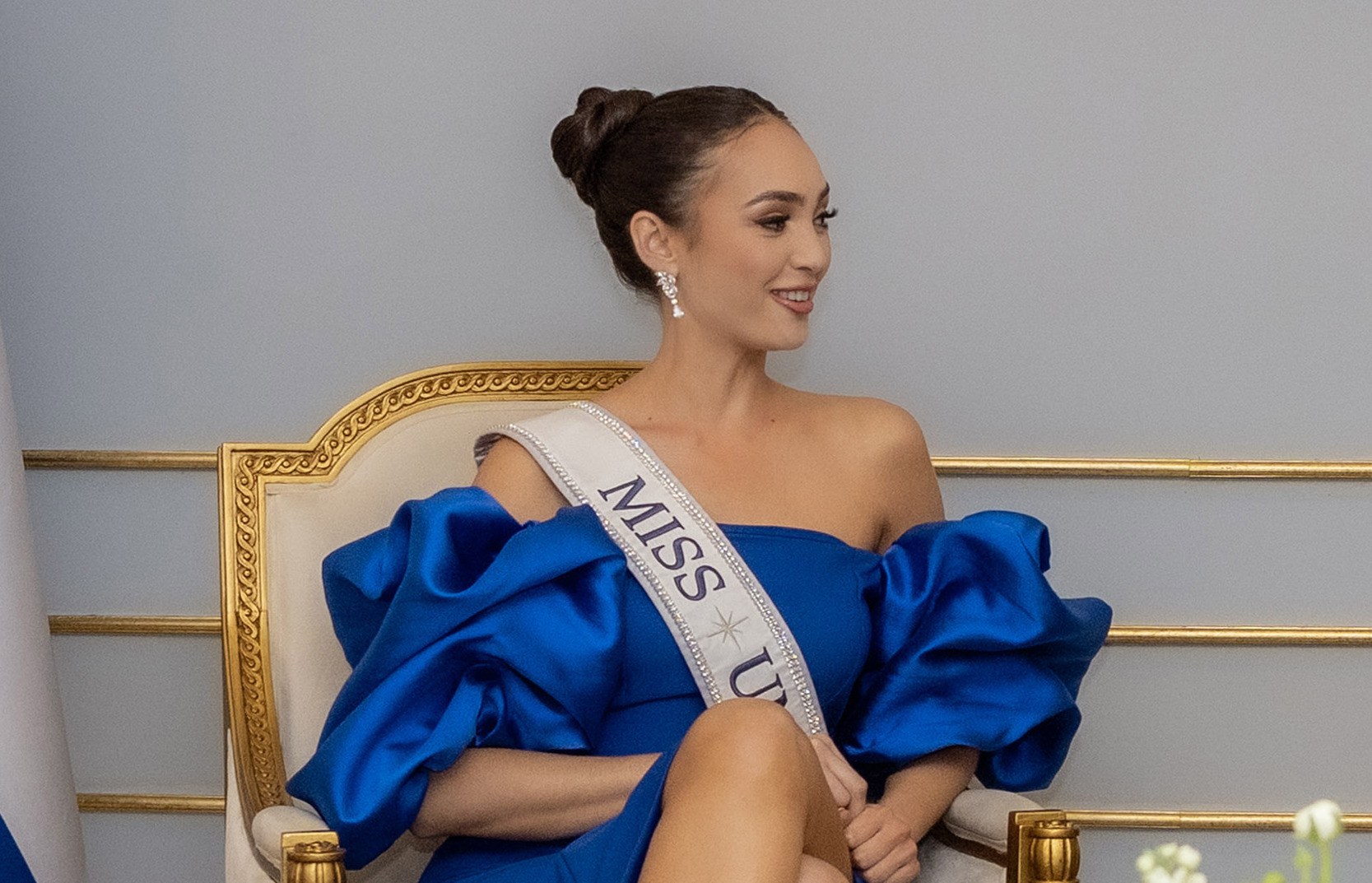 Frau in einem blauen Kleid mit einem "Miss Universe 2019"-Sash, die lächelt, während sie auf einem Stuhl sitzt, umgeben von Blumen mit einer Wand im Hintergrund.