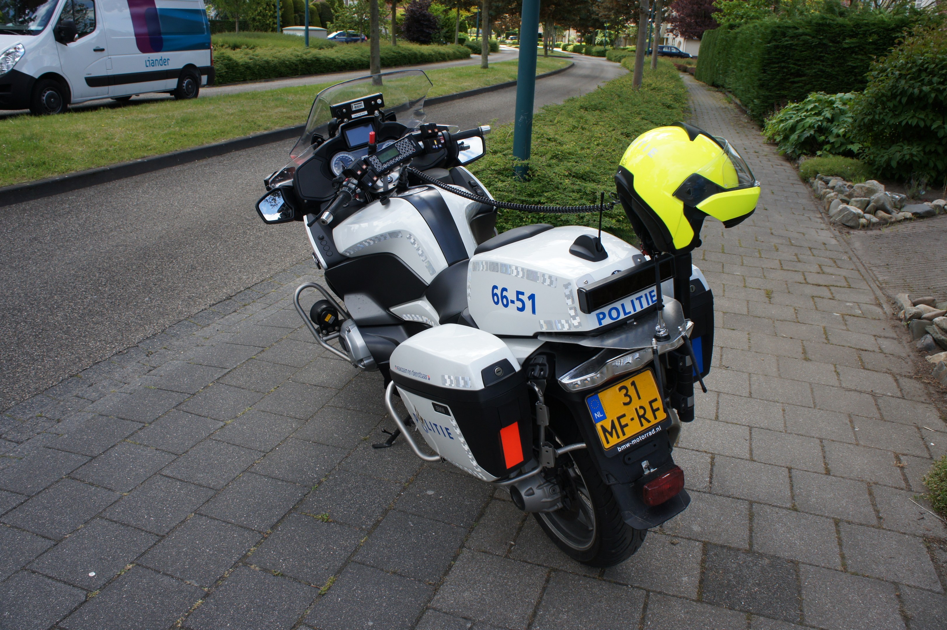 Polizeimotorrad am Straßenrand geparkt mit einem Helm darauf, umgeben von Bäumen, Pfählen, Gebäuden, Pflanzen, Gras, Steinen und anderen Fahrzeugen im Hintergrund.