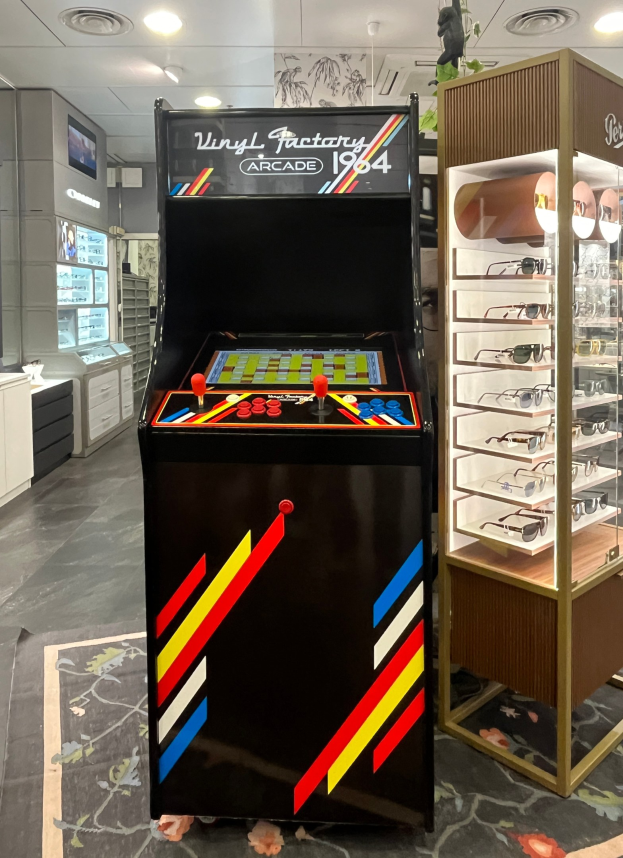 Ein Geschäft mit einem zentralen Retro-Arcade-Automaten, einem Gestell mit Gläsern auf der rechten Seite und Hintergrundelementen wie Schränken, einem Fernseher und anderen Gegenständen, wobei Deckenlampen eine retrohafte Atmosphäre schaffen.