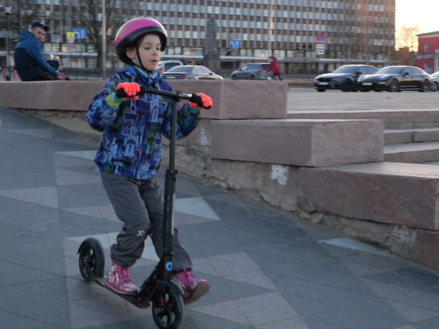 Ein junger Junge in Helm und Handschuhen fährt einen Roller eine Treppe hinunter, vorbei an Fahrzeugen, Menschen, Bäumen, Pfählen, Brettern, Gebäuden und einem klaren blauen Himmel.