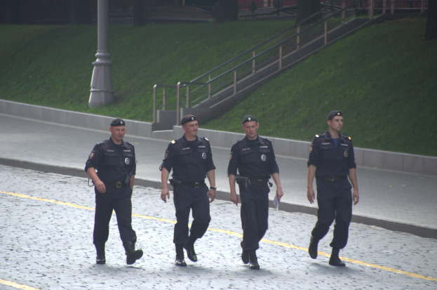 Vier Polizisten in Uniform gehen eine Straße entlang, jeder hält Papiere, mit einem Pfahl, Treppe, Geländer, Gras, Bäumen und einem Gebäude im Hintergrund.