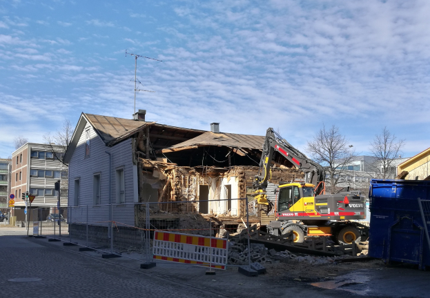 Baustelle mit einem großen Gebäude, das abgerissen wird, mit einem Kran, Zaun, Absperrung, Steinen, Container, Bäumen, Pfosten, Hinweisschild, Straßenlaterne, Fahrzeugen, Gebäuden und einem bewölkten Himmel.