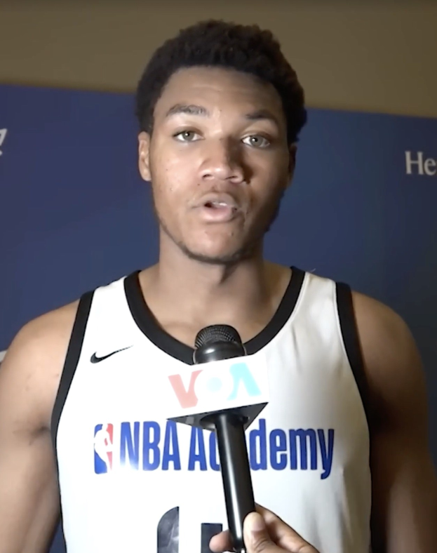 Mann in weißem Tanktop mit Mikrofon, spricht mit Medien vor "NBA All-Star Game 2019"-Plakat.