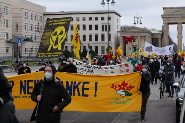 Eine große Gruppe von Menschen marschiert mit Transparenten und Fahnen auf einer Straße bei einer Demonstration gegen Atomkraft in Deutschland, mit Fahrzeugen auf der rechten Seite, Gebäuden und einem Bogen im Hintergrund und Bäumen, die die Straße säumen.