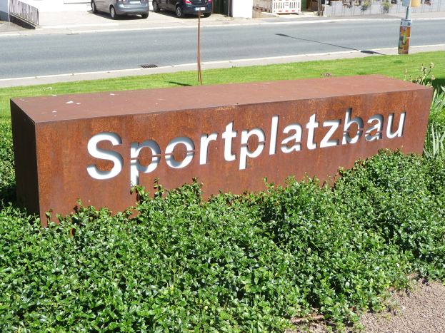 Ein Schild mit der Aufschrift 'Sportplatzbau' steht vor einem Gebäude, umgeben von grünem Gras und Pflanzen, mit Fahrzeugen, Gebäuden, Polen, Brettern und anderen Gegenständen im Hintergrund.
