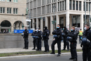 Polizisten in schwarzen Uniformen und Masken stehen vor einem Gebäude mit Glasfenstern und Säulen in Berlin, Deutschland, einige halten Helme, mit Laternen, Verkehrsampeln, Schildern mit Text, einer Statue und anderen Menschen in der Szene.