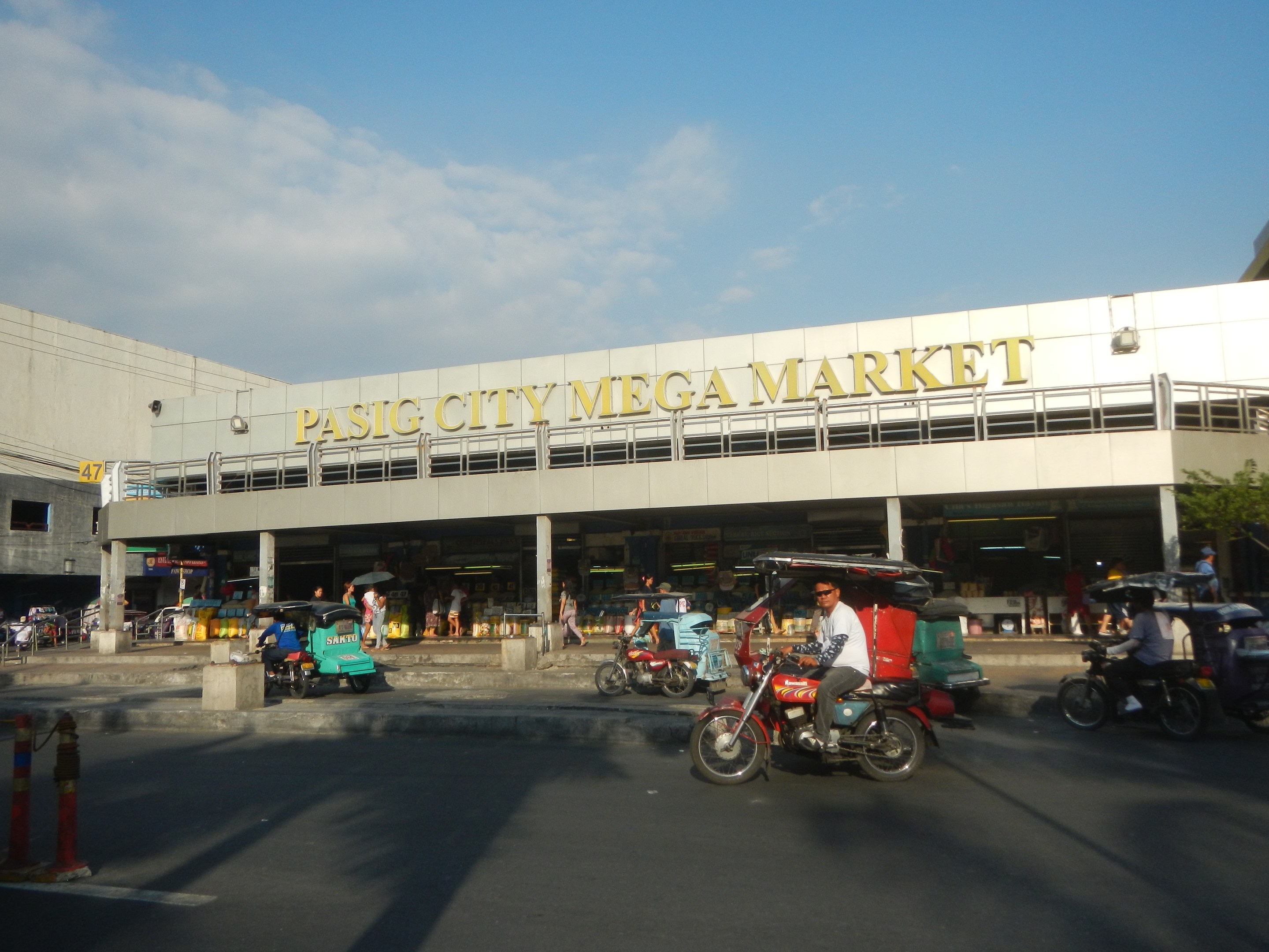 Eine belebte Straßenszene auf dem Pasig City Mega Market mit Fahrzeugen und Fußgängern, einem Gebäude mit Text im Hintergrund und einem bewölkten Himmel.