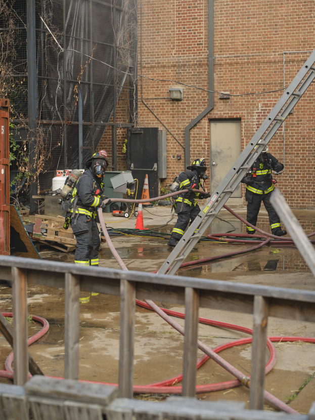 Feuerwehrleute in Helmen arbeiten daran, ein Gebäude Feuer zu löschen, während sie in der Nähe eines Metallzauns, verstreuter Rohre, eines Containers und eines Verkehrskegels stehen, mit einem Baum und Himmel im Hintergrund.