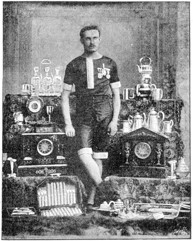 Schwarzes und weißes Foto eines Mannes in einem Fußballtrikot, der vor einem Tisch mit Pokalen und Trophäen steht, mit der Aufschrift "1909-1910 WM-Sieger" unten.