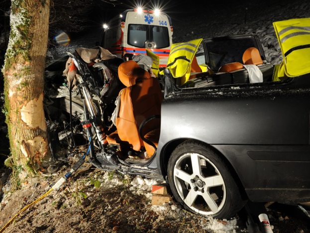 Ein beschädigtes Auto am Straßenrand bei Schnee mit einem im Hintergrund sichtbaren Rettungswagen, umgeben von Bäumen.