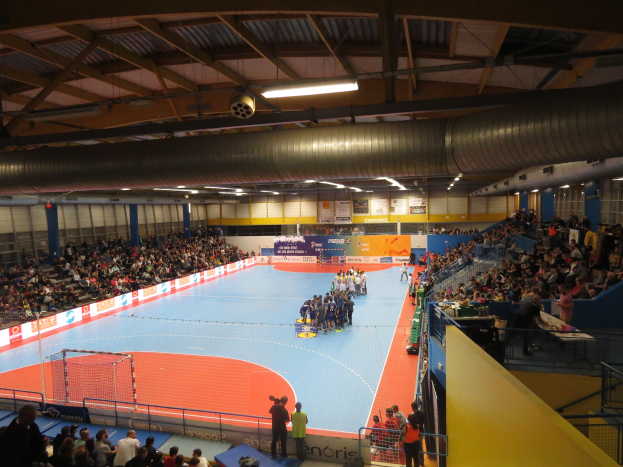 Große Halle mit Zuschauern bei einem Handballspiel, mit Sitzplätzen und Stehplätzen, Netz auf der linken Seite, Hintergrundtafeln mit Text und Deckenbeleuchtung und Rohre.