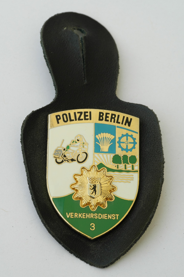 Ein schildförmiges Polizeimarke mit "Polizei Berlin" in schwarzer fetter Schrift auf einem blauen Hintergrund mit einem weißen Stern und Rahmen, das auf einer Oberfläche ruht.