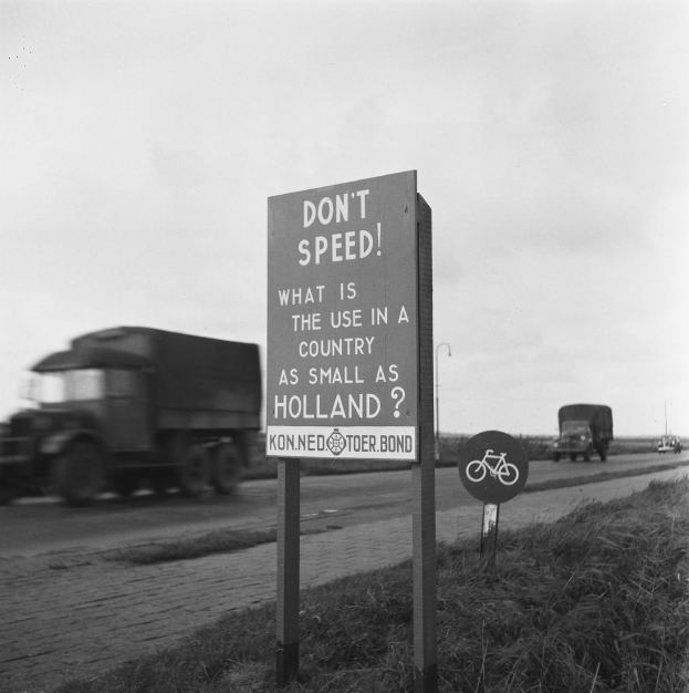 Schwarz-weißes Foto eines Lkw, der auf einer Straße mit einem "Fahr nicht zu schnell"-Schild im Vordergrund und anderen Fahrzeugen, Gras, Masten und einem klaren Himmel im Hintergrund fährt.